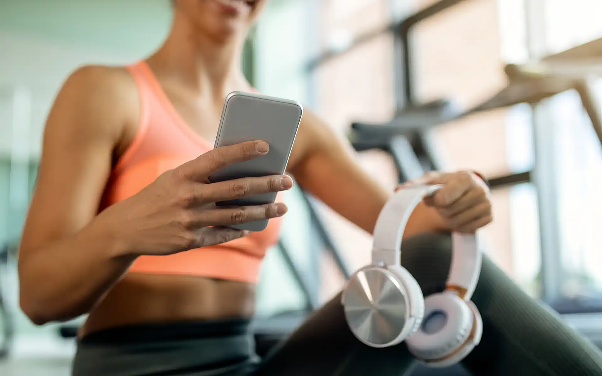 A woman holding headphones in a gym looking at her phone using the Project U App